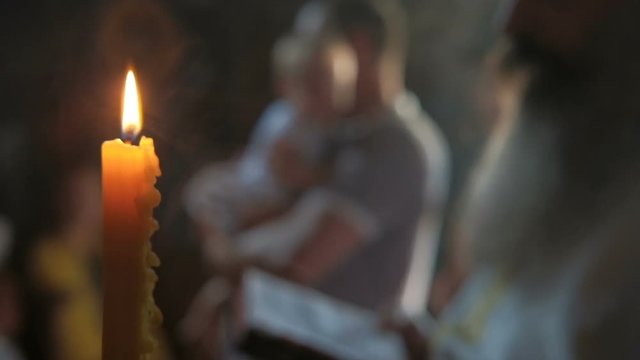 Lit candle on background of father with child ready for church ritual. Behind flame man holds a child in his arms for the rite of baptism in church.