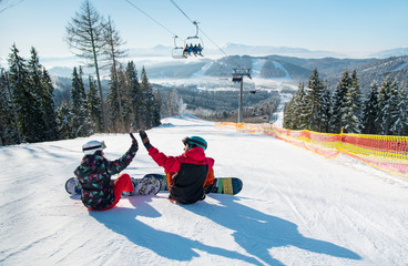 Snowboarders sit on the top of the ski slope under the ski lift let's high five to each other with a beautiful scenery of mountains and forests on a sunny morning
