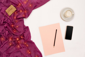 A cup of coffee, black smart, pink paper with new years goals. Scarf and Christmas lights on white background.