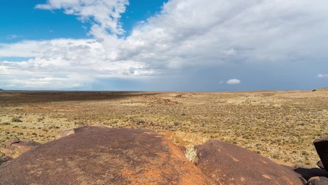 A linear timelapse on a bright sunny day framed with rocks in the foreground of a rocky Karoo landscape with wide open vistas and dramatic stormy clouds gathering while rain is falling in the distance. 