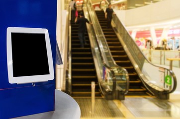 blank screen digital monitor or tablet on counter in front of escalator in modern department store shopping mall, copy space for text or media content, marketing and advertisement concept