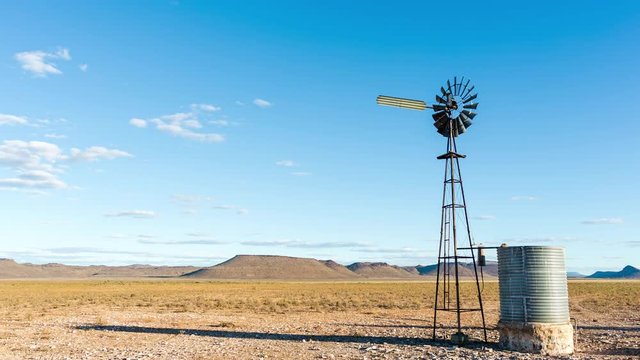 A slow linear day to night time-lapse of a typical arid Karoo farm landscape, a windmill blowing in the wind with scattered cumulous clouds against a bright blue sky as the night falls into a moonlit landscape and sky filled with stars. 