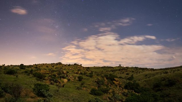 A linear fast motion timelapse at night of a bright moonlit landscape and abstract rocks in the foreground revealing a lush green, rocky landscape with clouds passing by. 