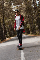 A young hipster in a cap and plaid shirt is riding his longboard on a country road in the forest