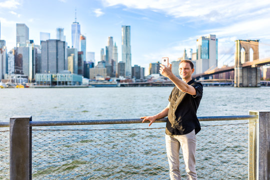Young hipster man standing taking selfie picture by fence in Brooklyn Bridge Park overlooking the NYC New York City Manhattan cityscape skyline with water bay during sunset