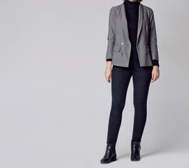 Woman wearing stylish and smart outfit with black turtleneck, gray elegant double-breasted blazer, black skinny jeans and black ankle boots isolated on gray background. Copy space