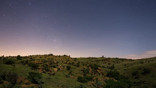 A linear fast motion timelapse at night of a bright moonlit landscape and abstract rocks in the foreground revealing a lush green, rocky landscape with shooting stars. 