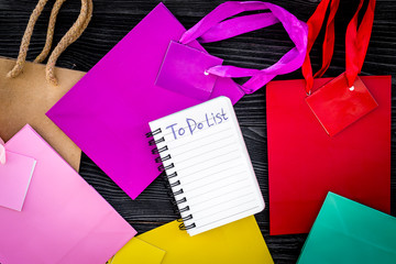 To do list for shopping among colorful paper bags on grey wooden background top view