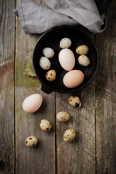 Whole quail and chicken eggs in a small cast-iron frying pan on the background of an old wooden texture. Selective focus. Top view with space