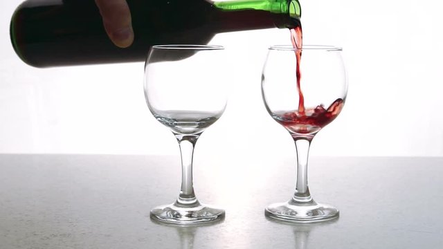 The two cristall glasses is being fulfilled with the red dry wine with rich bouquette in the restaurant bar. The liquid is pouring slowly from the green bottle and waving and smashing inside the