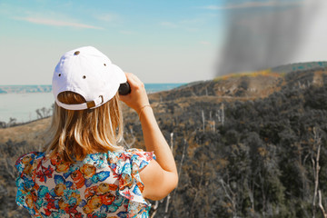 A girl in a blouse with flowers and a white hat looks through binoculars at a burning and smoking...