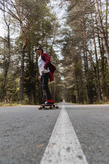 A young hipster in a cap and plaid shirt is riding his longboard on a country road in the forest