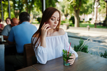 Portrait of an attractive young businesswoman holding a mojito cocktail and talking on the phone in a cozy cafe next to the park.