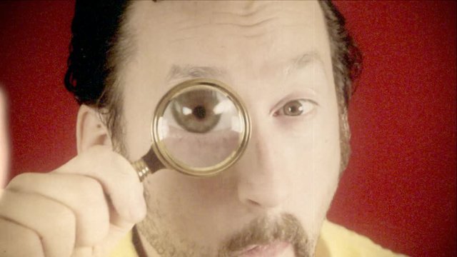 Fake 8mm amateur film: a funny ugly man looking at the camera with a magnifying lens (a detective / private eye), searching for valued information.
