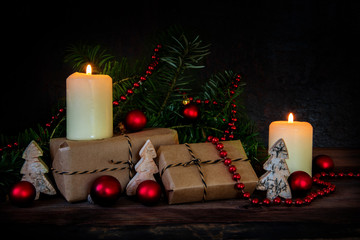 gift boxes in kraft paper and two burning candles decorated with red christmas baubles, fir branches and small wooden trees on a dark rustic background with copy space