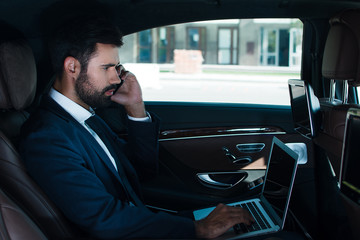 On his way to office. Side view of handsome young man using laptop and talking on mobile phone while sitting in car