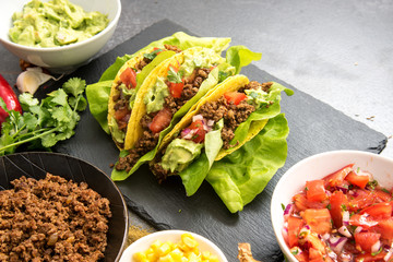 mexican tacos and some ingredients like fried ground beef, tomato salsa, guacamole, corn and herbs on a dark slate plate with copy space