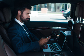 Checking emails on a go. Side view of handsome young man using laptop and using his smart phone while sitting in car