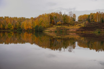 The bank of the river is reflected in the mirror calm water in the autumn cloudy day.