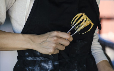 Woman baking in black apron, holding beaters covered in batter. 