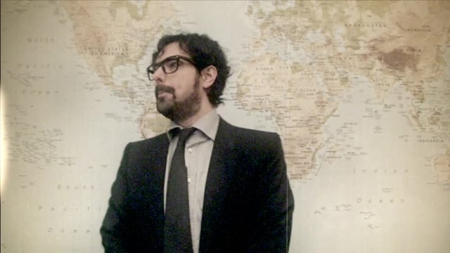 Fake 8mm amateur film: a man in an elegant suit showing a bit of funny madness over a world map, possibly suggesting a broadcast weather report or a teacher teaching in a geography class.

