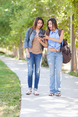 Obraz premium Two Beautiful Young Ethnic Twin Sisters With Backpacks Using A Smartphone Outside.