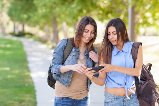 Two Beautiful Young Ethnic Twin Sisters With Backpacks Using A Smartphone Outside.