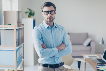 Portrait of strict serious smart successful boss dressed in blue shirt and trousers. He is standing with crossed arms in his workstation