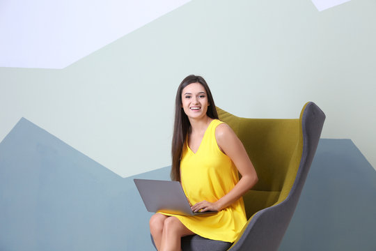Young woman with laptop in armchair against color background