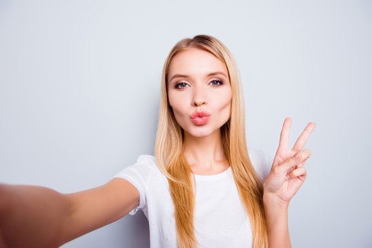 Beautiful young pretty woman with straight blonde hair sending air kiss and showing victory sign to her boyfriend via video connection. She is isolated on grey background