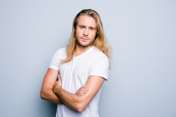 Close up portrait of serious and confident young guy with bristle and long blonde hair, he is standing with crossed arms over grey shadeless background