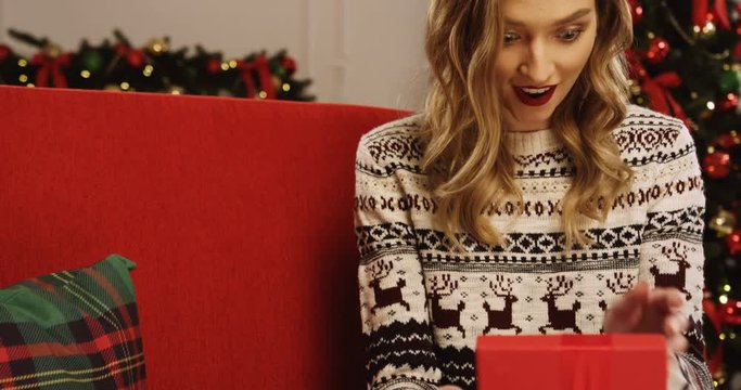 Blonde young woman opening Christmas present with red ribbon on it with surprised face while sitting on the red couch in the living room on the Christmas tree with light background. Indoor. Close up