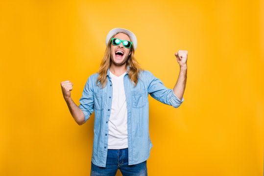 Yes! Lucky young hipster man isolated over yellow background, happy man wearing glasses and hat acts like he won something with raised hands, emotion, success, gesture and people concept