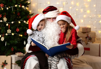 Fototapeta na wymiar Santa Claus reading magic book to happy little cute children boy and girl kids near Christmas tree