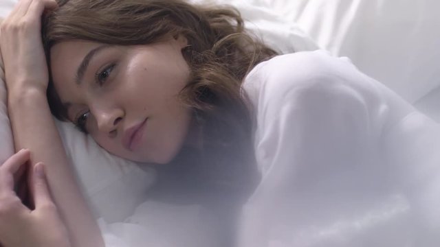 Slowmo of elegant young woman in nightgown lying in bed and looking around sleepily while waking up in morning