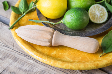 Limes with a lemon on a yellow plate and a wooden table, next to a juicer for citrus.
