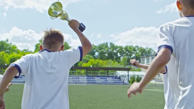 Handheld tracking of happy boys from junior soccer league holding gold cup and celebrating while running across playing field with green grass