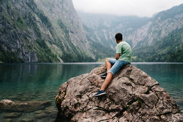Young adult man sitting on rock looking at beautiful and misty l