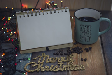 The golden inscription Marry Christmas, a blue cup and coffee beans, an open notepad for entries and glowing New Year's lights on the table