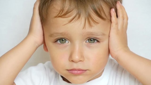 A close-up of a child looks thoughtfully at the camera, holding his hands behind his head, closing his ears. The child is thoughtful, does not want to hear anyone
