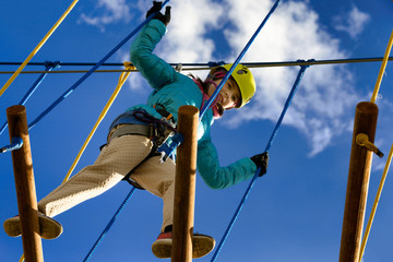Happy girl in a yellow helmet and a blue jacket is playing in an adventure park holding ropes and climbing a wooden and rope staircase. Bottom view.