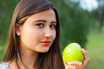 Beautiful teenage girl biting an apple, against green of summer park.
