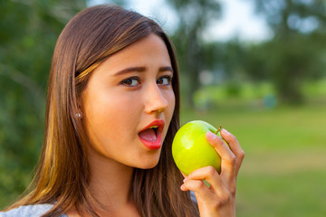 Beautiful teenage girl biting an apple, against green of summer park.