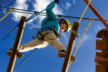 Happy girl in a yellow helmet and a blue jacket is playing in an adventure park holding ropes and climbing a wooden and rope staircase. Bottom view. © Slava
