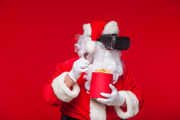 Santa Claus wearing virtual reality goggles and a red bucket with popcorn, on a red background. Christmas