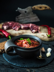 Borsch traditional Ukrainian and Russian soup in ceramic bowl with rye bread, chilly paper and garlic. Meat and butcher knife on the background.