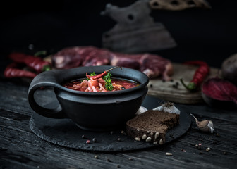 Borsch traditional Ukrainian and Russian soup in ceramic bowl with rye bread, chilly paper and garlic. Meat and butcher knife on the background.