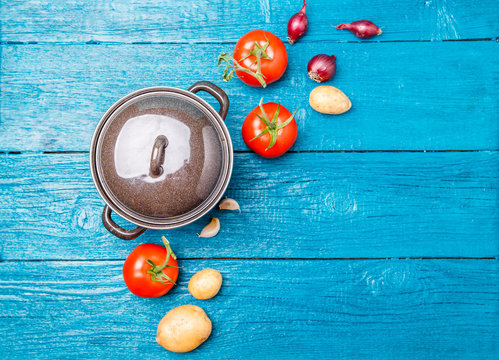 Photo on top of iron pot, tomato, potato, onion on blue wooden background.