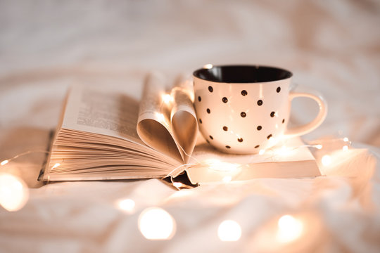 Open book with folded sheets in heart shape and cup of tea in bed with Christmas lights closeup. Good morning. Breakfast time.