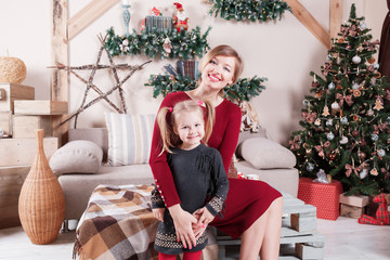 Obraz na płótnie Canvas Cute pregnant mom and daughter sitting near beautiful decorated Christmas tree at home, looking at camera, happy pregnancy time